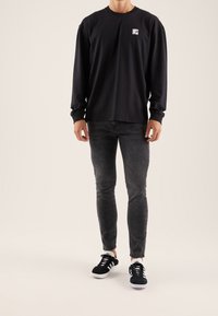 T-shirt nera a maniche lunghe con patch logo, abbinata a jeans skinny grigio scuro e scarpe nere con dettagli bianchi.
