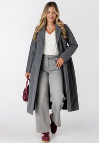 Vrouw die loopt terwijl ze een lange grijze jas, een wit gebreid topje, grijze wijde jeans, bordeauxrode sneakers en een kleine bordeauxrode handtas draagt.