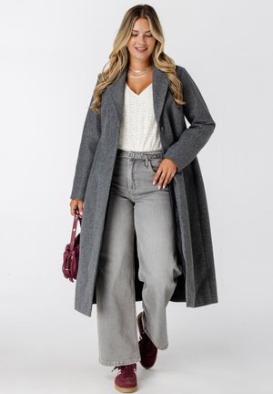 Vrouw die loopt terwijl ze een lange grijze jas, een wit gebreid topje, grijze wijde jeans, bordeauxrode sneakers en een kleine bordeauxrode handtas draagt.