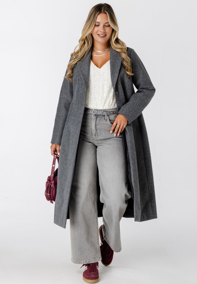 Vrouw die loopt terwijl ze een lange grijze jas, een wit gebreid topje, grijze wijde jeans, bordeauxrode sneakers en een kleine bordeauxrode handtas draagt.