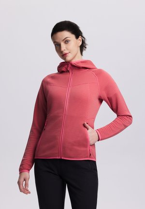 Woman with dark hair wears a pink zip-up hooded jacket with textured fabric, black pants, and poses with one hand in pocket against a plain background.