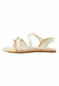 Sandalia blanca con múltiples correas, un acento de purpurina texturizada y una hebilla dorada. Suela plana con un patrón de relieve en marrón claro.