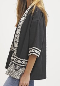 Kvinde iført en sort kimono-lignende jakke med hvid geometrisk broderi på ærmerne og kanten, parret med lyseblå jeans.