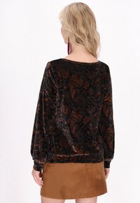 Schwarze Samtbluse mit Paisleymuster in Orange- und Brauntönen, mit langen Ärmeln und lockerer Passform, kombiniert mit einem braunen Wildlederrock.