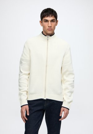 Uomo che indossa un maglione bianco con zip, con polsini e orlo a coste, in piedi davanti a uno sfondo chiaro e semplice.