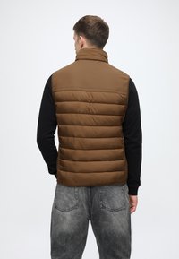 Barna pufferszövet mellény magas nyakkal, steppelt mintával, fekete hosszú ujjú pólóval párosítva. Szürke, megviselt farmernadrág látható.