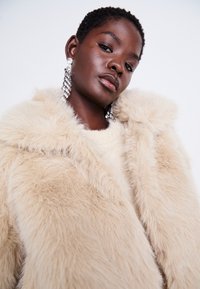Manteau en fausse fourrure beige avec un large col, présentant une texture douce et moelleuse. De grandes boucles d'oreilles en cristal complètent le look.