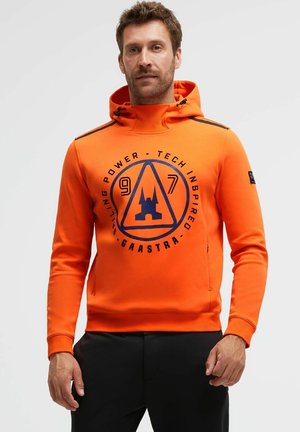 Man wearing bright orange hoodie with black graphic logo and text "Gaastra" on chest, paired with black pants, standing against plain background.