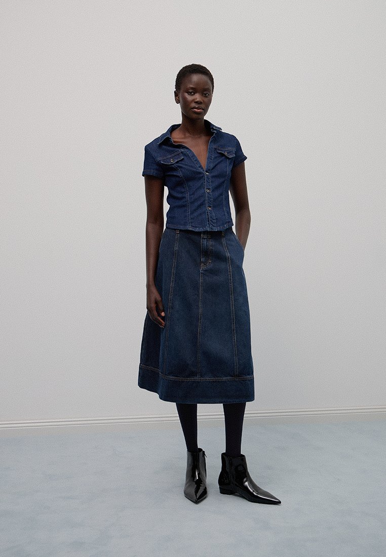 Dżinsowy komplet składający się z dopasowanej koszuli z krótkim rękawem i spódnicy do kolan. Oba elementy są w ciemnoniebieskim kolorze z widocznymi detalami ściegów.