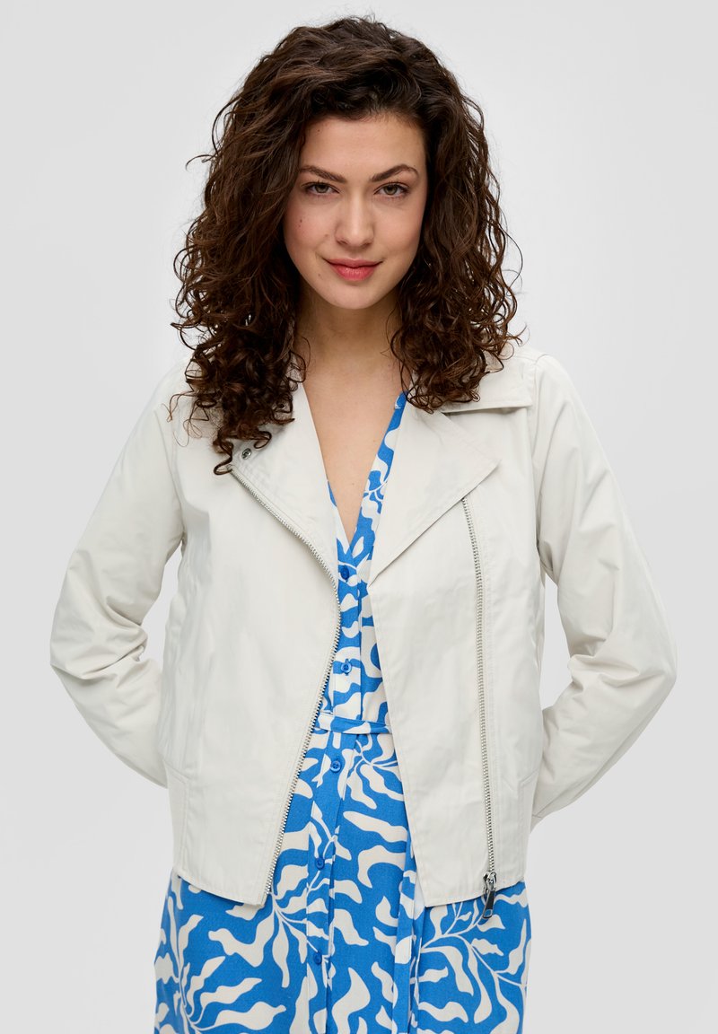 Lichte witte jas met rits aan de voorkant, met een afgeronde kraag en lange mouwen. Draagt over een blauw-wit patroonjurk.