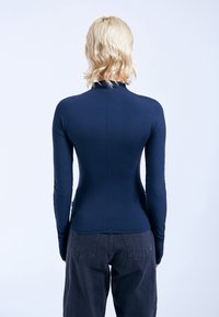 Marineblaue Langarmbluse mit hohem Kragen, figurbetontem Design und weicher Textur. Kombiniert mit dunkelgrauen Hosen mit Gesäßtaschen.