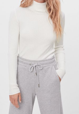 Woman wearing a white ribbed turtleneck sweater and light gray drawstring sweatpants, with one hand in pocket and loose hair.