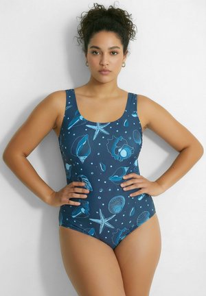 Mujer con cabello rizado que lleva un traje de baño de una pieza azul con patrones de conchas marinas y estrellas de mar en tonos azul claro, de pie con las manos en las caderas.