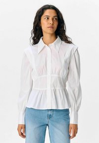 Witte overhemel met schouderplooien, een aansluitend lijfje en wijdvallende mouwen. Draagt met lichtblauwe jeans. Glad, lichtgewicht stof.