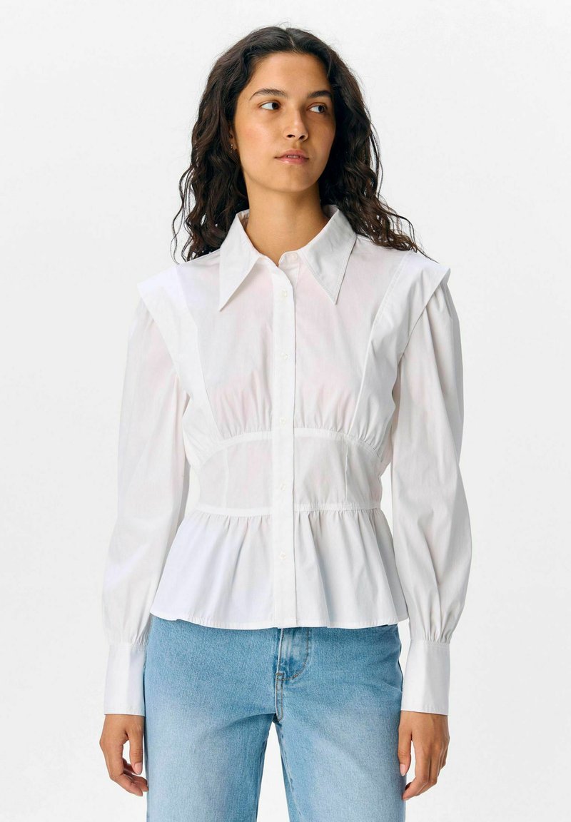 Witte overhemel met schouderplooien, een aansluitend lijfje en wijdvallende mouwen. Draagt met lichtblauwe jeans. Glad, lichtgewicht stof.