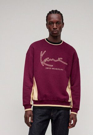 Mladenič s pletenicami, ki nosi bordno-bež pulover z napisom "Kang 1989 EST. BROOKLYN, NYC", stoji pred enobarvnim ozadjem.
