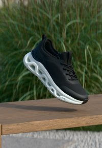 Μαύρο αθλητικό παπούτσι με λευκή σόλα που διαθέτει γεωμετρικές εγκοπές, εμφανίζεται στον αέρα πάνω από ξύλινη επιφάνεια με πράσινα φυτά στο φόντο.