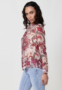 Camicetta trasparente con un vivace motivo paisley in tonalità di rosso e rosa, caratterizzata da un colletto e maniche lunghe, abbinata a jeans chiari.