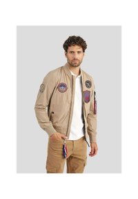 Veste bomber beige avec une fermeture éclair à l'avant, des poignets côtelés et plusieurs écussons sur les manches. Associée à un pantalon cargo beige et un t-shirt blanc.