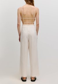 Rückansicht eines hellbeigen Jumpsuits mit dünnen Trägern, offenem Rückendesign und weitem Bein. Verfügt über eine glatte Textur und einen weichen Fall.