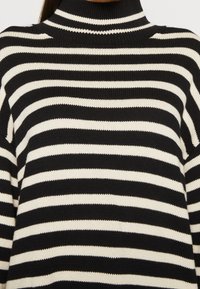 Pull en maille à col roulé rayé horizontalement noir et blanc porté par une personne, montrant le haut du torse et le cou.