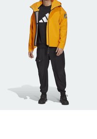 Gelbe wasserdichte Jacke mit Kapuze, grauen Akzenten und einem Logo. Kombiniert mit schwarzen Cargo-Hosen und schwarzen Schuhen, steht vor einem hellen Hintergrund.