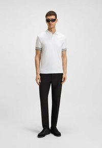 Vit polo med grårandiga manschetter, kombinerad med svarta byxor och svarta sneakers; modell som bär mörka solglasögon, står mot en neutral bakgrund.