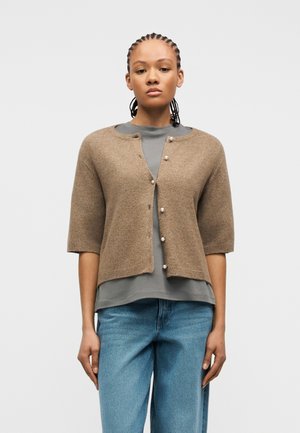 Vrouw met gevlochten haar, gekleed in een bruine korte mouw cardigan met parelknopen over een grijs shirt en een blauwe spijkerbroek, staand tegen een effen achtergrond.