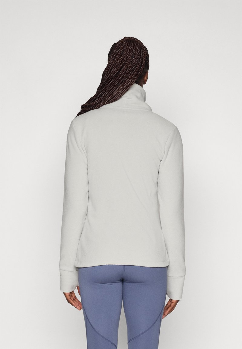 Lichtgrijze fleecejack met hoge kraag, zachte textuur en lange mouwen; gedragen met blauwe sportlegging.
