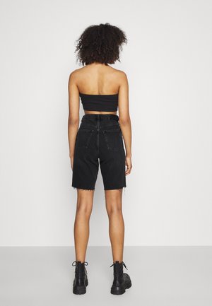 Person mit lockigem Haar, die ein schwarzes Tube-Top, hoch taillierte schwarze Denim-Shorts und schwarze Schnürstiefel trägt, sieht man von hinten auf einem einfarbigen Hintergrund.