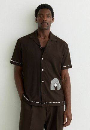 Mees, kes kannab pruuni lühikeste varrukatega särki, mille varrukatel ja äärel on valged lainelised jooned ning vöökohal valge abstraktne muster.