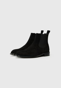 Bottines Chelsea en daim noir avec panneaux élastiques sur les côtés, bout rond et semelle noire fine. Comprend des languettes pour un enfilage facile.