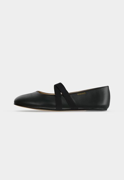 Chaussure de ballet plate en cuir noir avec des bandes élastiques croisées et le logo Versace sur le côté intérieur, vue de profil sur fond blanc.