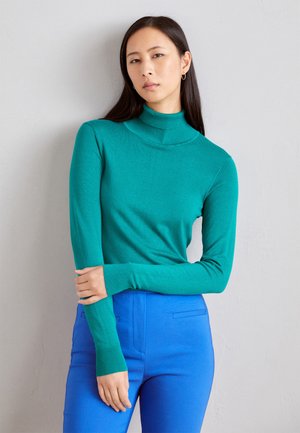Pull à col montant teal en tricot doux, avec des manches longues et des poignets côtelés. Associe à un pantalon bleu ajusté.