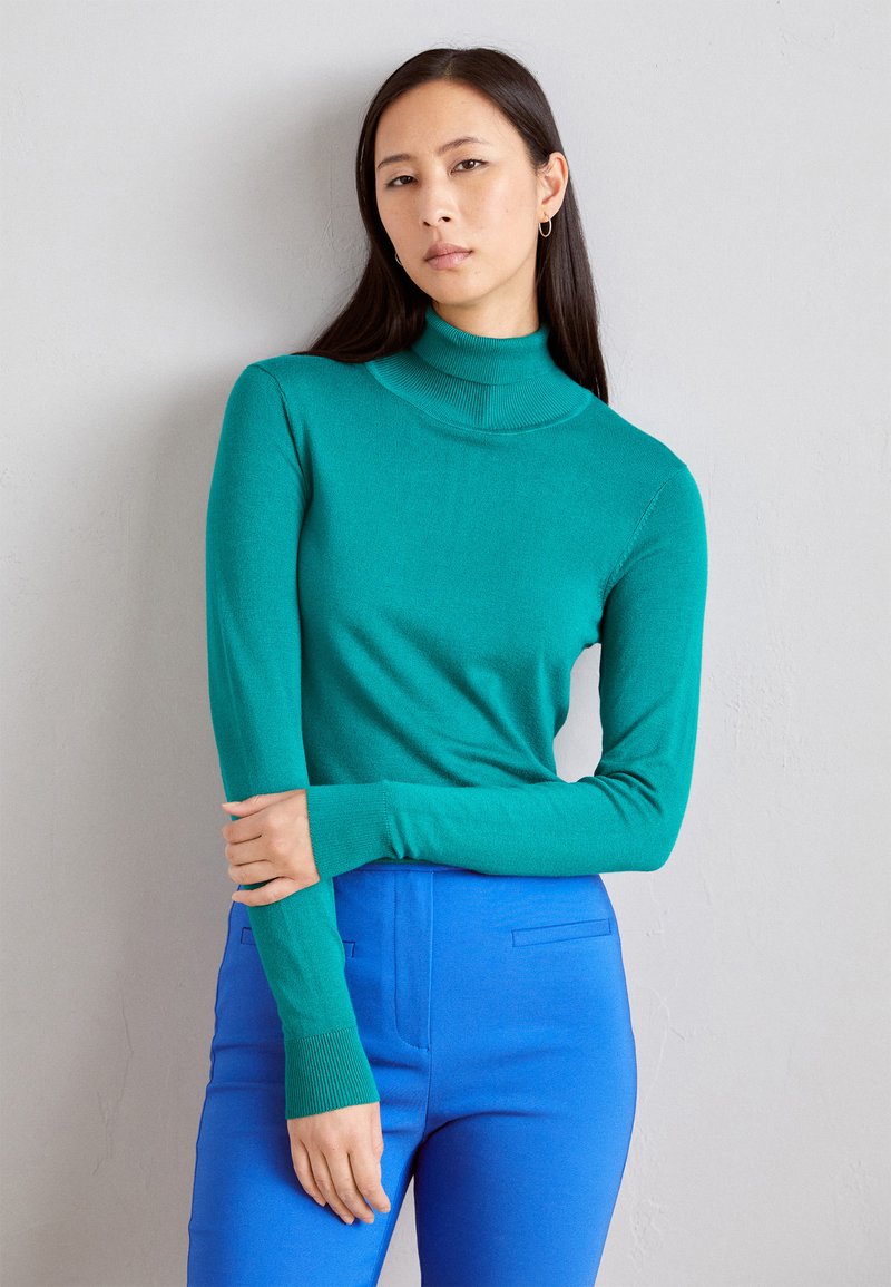 En teal turtleneck sweater lavet af blødt strikket stof, med lange ærmer og ribbede manchetter. Sammen med blå tætsiddende bukser.