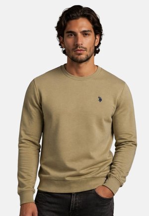 Mann mit dunklem Haar trägt beige Rundhalspullover mit kleinem dunkelblauem Polo-Spieler-Logo, steht mit einer Hand in der Tasche vor einfarbigem Hintergrund.