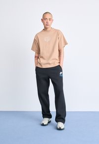 Bež majica kratkih rukava s bijelim logotipom, uparena s crnim treniranim hlačama koje imaju mali plavi logotip. Obučena s teksturiranim bijelim tenisicama.