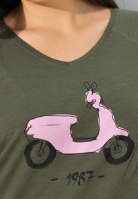 Oliwkowy T-shirt z grafiką różowego skuteru z czarnymi konturami, z towarzyszącym rokiem „1987” w wyraźnej czcionce poniżej.