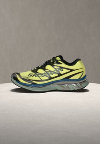 Κίτρινο και μαύρο αθλητικό παπούτσι με πάχυ, κυρτή σόλα και μπλε λεπτομέρεια, τοποθετημένο πλαγίως σε φωτεινή επιφάνεια με απλό φόντο.