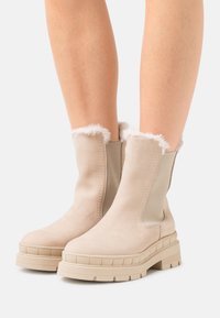 Beige ankle boots with a smooth suede finish, rounded toe, rubber sole, and faux fur trim at the top. Features elastic side panels.
