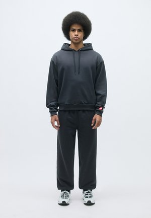 Tumši pelēki kapuci un atbilstoši sporta bikses, abi izgatavoti no mīksta auduma; kapucei ir aukla, un biksēm ir brīvs piegriezums. Baltas sporta kurpes.