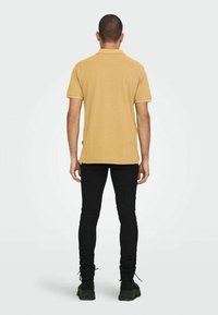 Polo de manga corta en color mostaza, hecho de tela texturizada, con cuello y tapeta, combinado con jeans ajustados negros y zapatos oscuros.