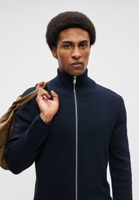 Μπλέ πλεκτό ζακέτα με φερμουάρ και ψηλό λαιμό, με μεταλλικό φερμουάρ. Το μοντέλο κρατά μια καφέ τσάντα στον έναν ώμο.