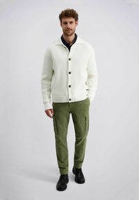 Weiße, gerippte Strickjacke mit schwarzen Knöpfen, kombiniert mit grünen Cordura-Cargo-Hosen mit Seitentaschen und schwarzem Schuhwerk.