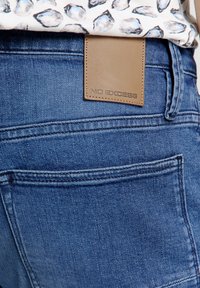 Blauwe denim jeans hebben een gladde textuur en een tan leren label met het reliëf "NO EXCESS" logo. De achterzakken zijn zichtbaar, met subtiele stiksels.