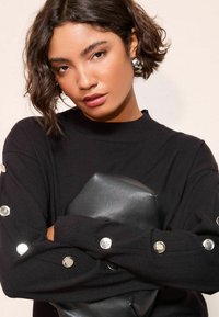 Pulover negru tricotat cu accente de nasturi argintii pe mâneci, având un guler striat. Ținând o geantă clutch mată de culoare neagră.