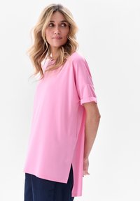 Różowa bluzka z krótkim rękawem o luźnym kroju, okrągłym dekolcie, bocznymi rozcięciami oraz gładkim materiale. Nosi się ją z ciemnoniebieskimi spodniami.