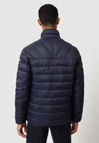 Uomo che indossa un giubbotto trapuntato blu navy, girato di spalle, in piedi contro uno sfondo chiaro e semplice.