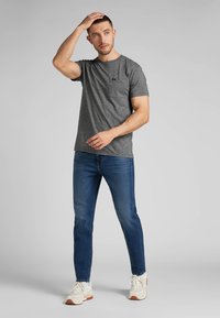 Grå kortærmet t-shirt med en brystlomme, parret med blå jeans og hvide sneakers. Stoffet ser blødt ud med en afslappet pasform.