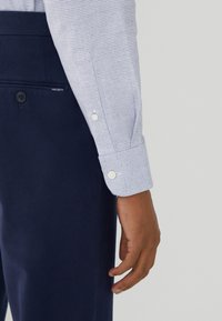 Camicia a quadri azzurro chiaro con bottoni bianchi, abbinata a pantaloni navy con tasca posteriore e etichetta con logo. Tessuto morbido.